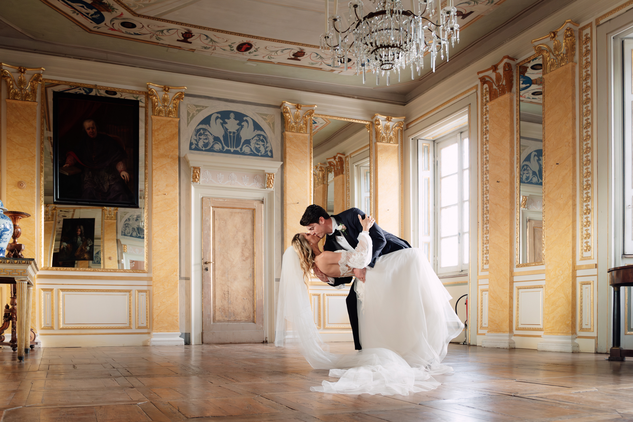Beatrice e Conrad danzano nel salone decorato, tra luce calda e dettagli dorati.