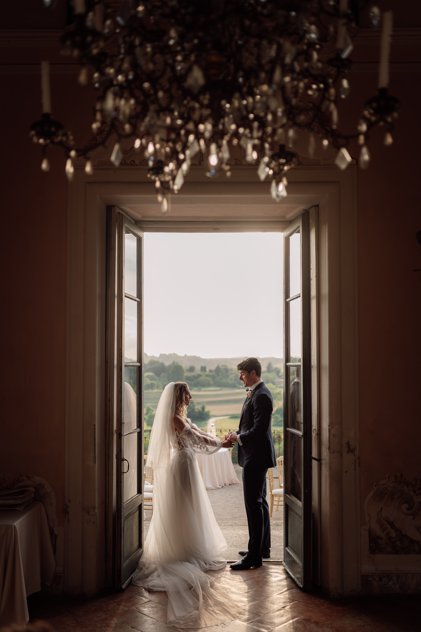 Beatrice e Conrad si tengono per mano davanti alla finestra illuminata dal tramonto.