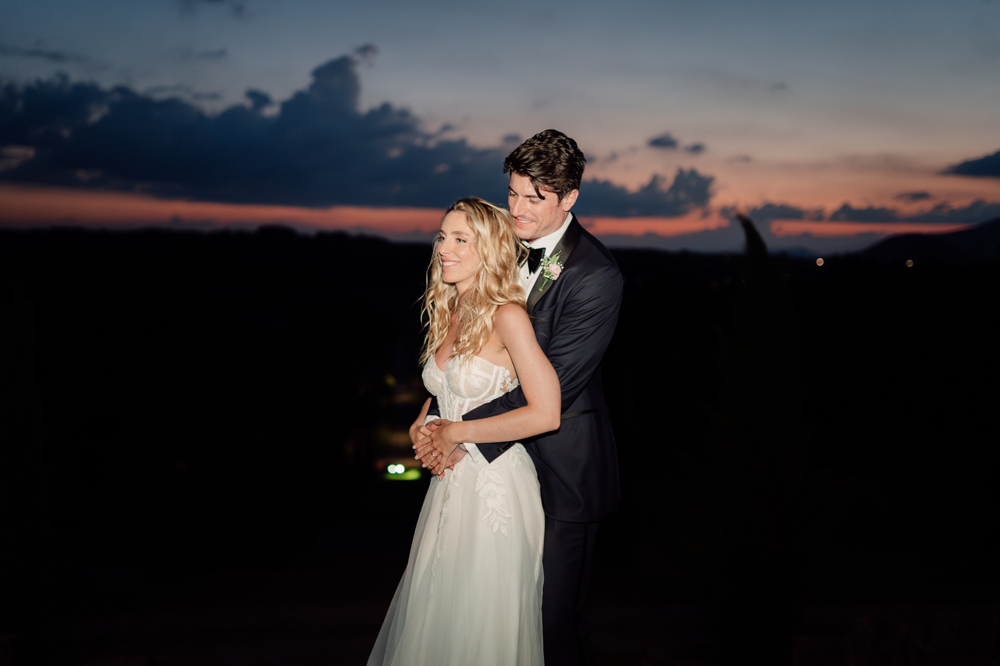 Lo sposo abbraccia la sposa con il tramonto sullo sfondo.