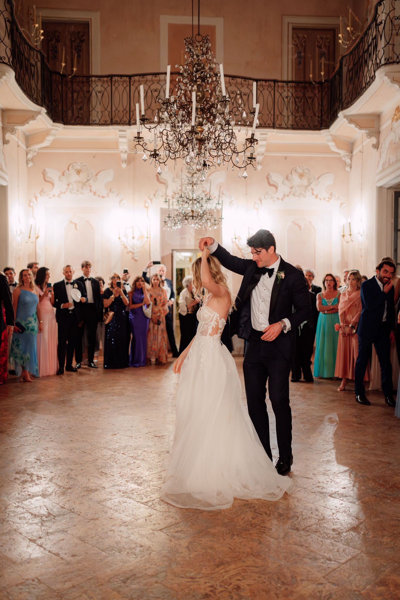 Gli sposi danzano al centro della sala, circondati dagli ospiti e dai lampadari scintillanti.