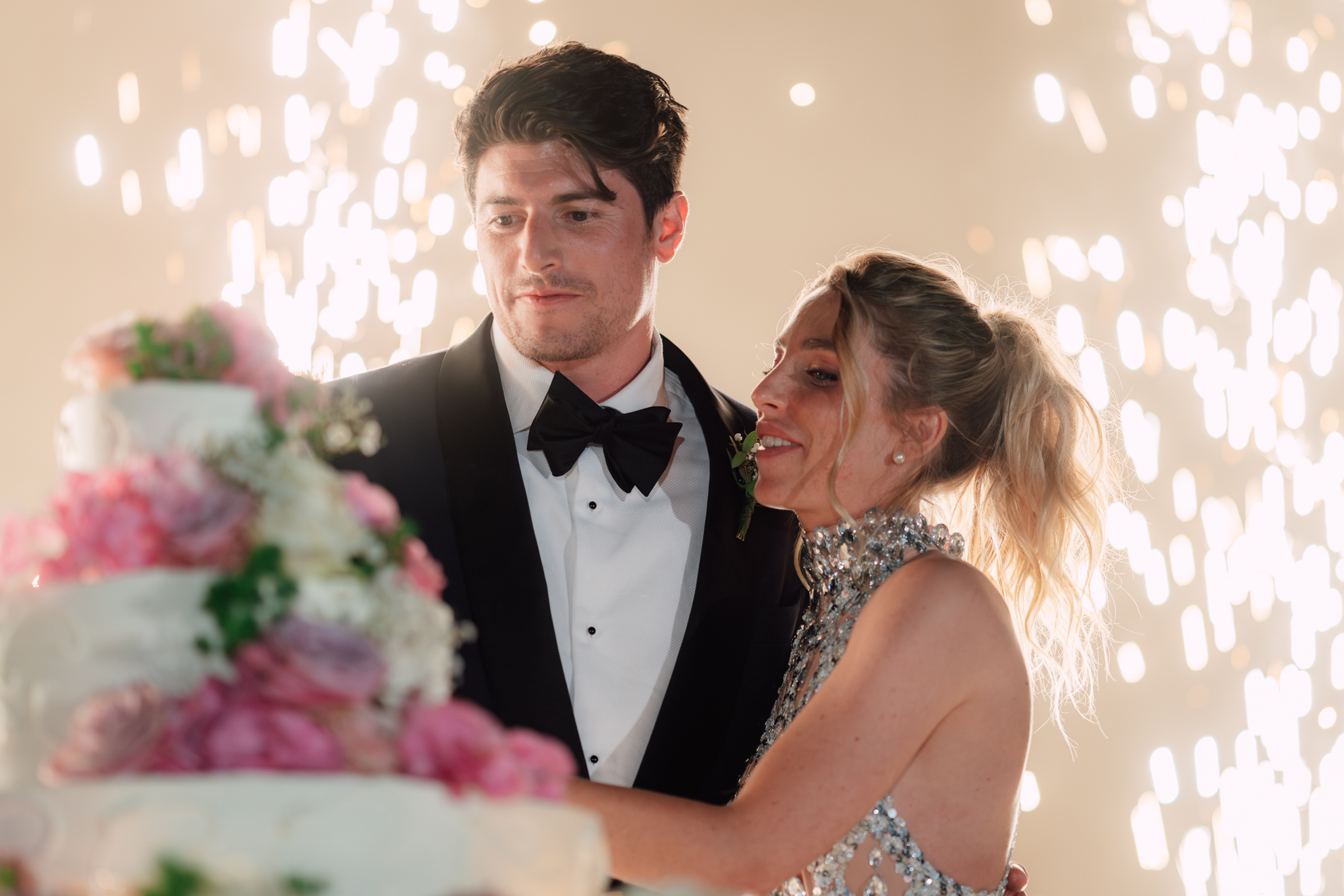 Gli sposi tagliano la torta tra giochi di luce e fiori rosa durante il ricevimento serale.