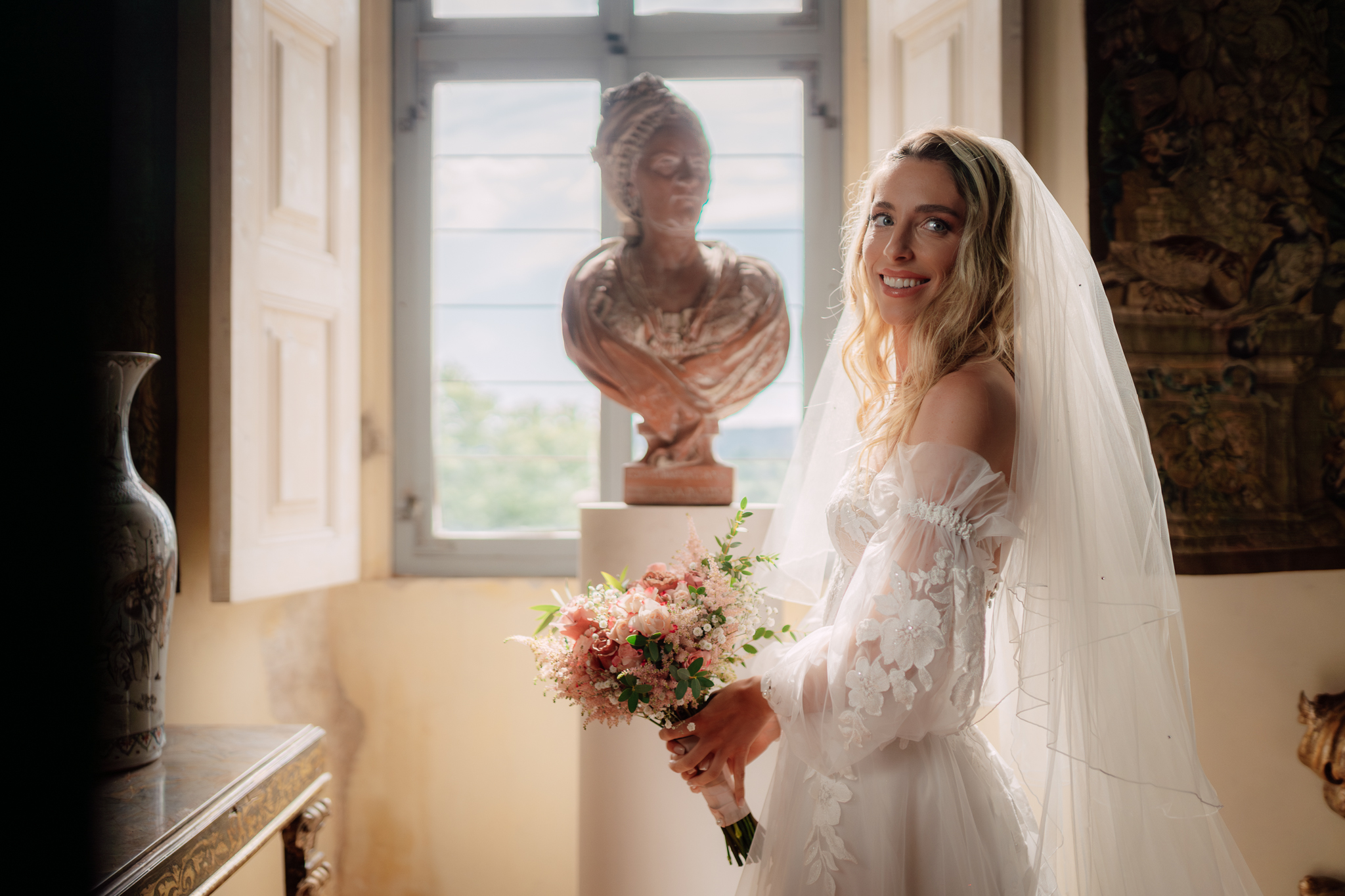 La sposa sorride accanto alla finestra con bouquet floreale, avvolta in luce naturale e romanticismo.